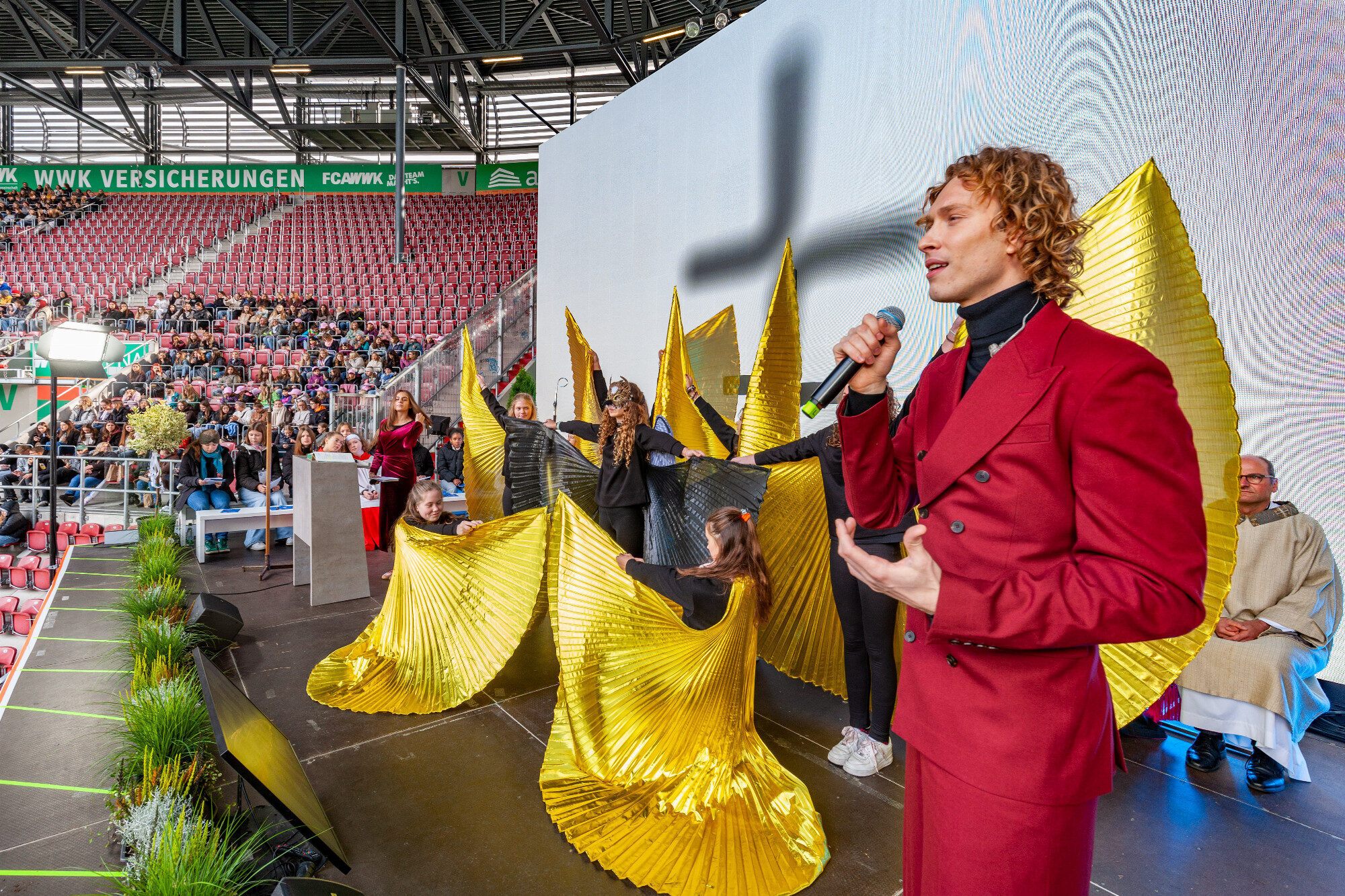 Sänger im bordeauxroten Anzug vor goldenen Flügeln, WWK Arena Augsburg / Schulwerk