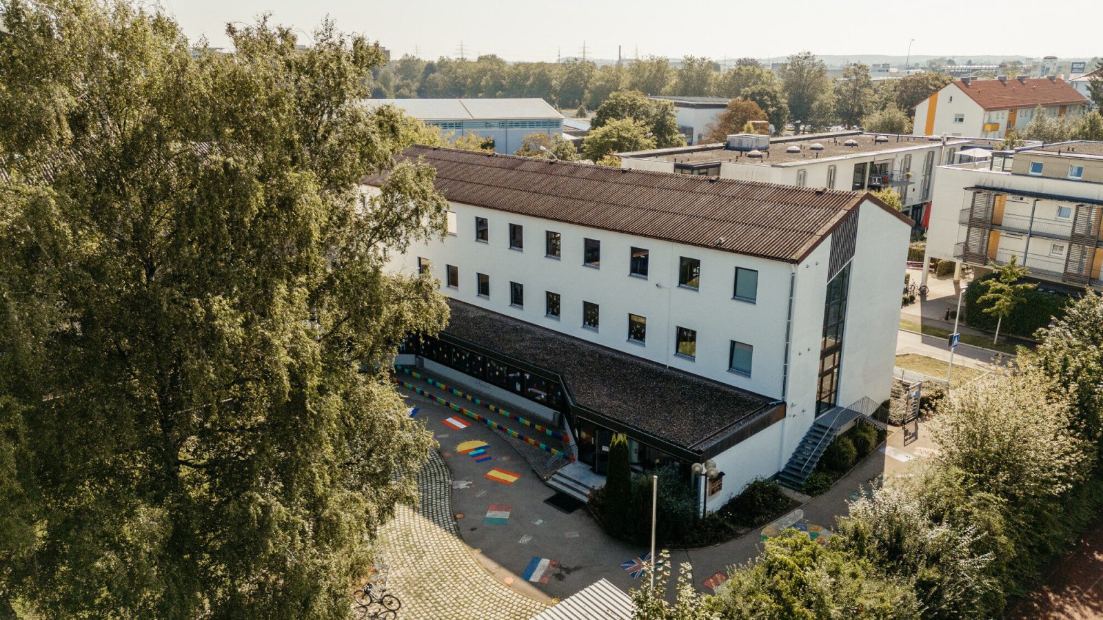Luftaufnahme des weißen Schulgebäudes der St.-Michael-Grundschule in Neu-Ulm mit buntem Pausenhof